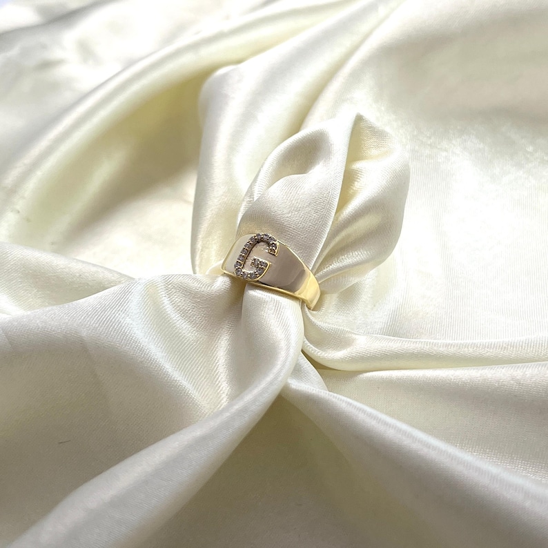 May include: A gold-colored signet ring with the letter "G" encrusted with small, clear gemstones. The ring is set against a backdrop of ivory-colored, silky fabric, creating a luxurious aesthetic. The ring's design is simple yet elegant.