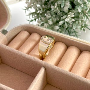 May include: A gold-colored ring with a rectangular face, adorned with small, clear gemstones forming a letter "G". The ring is set against a light pink jewelry box with a white floral background.