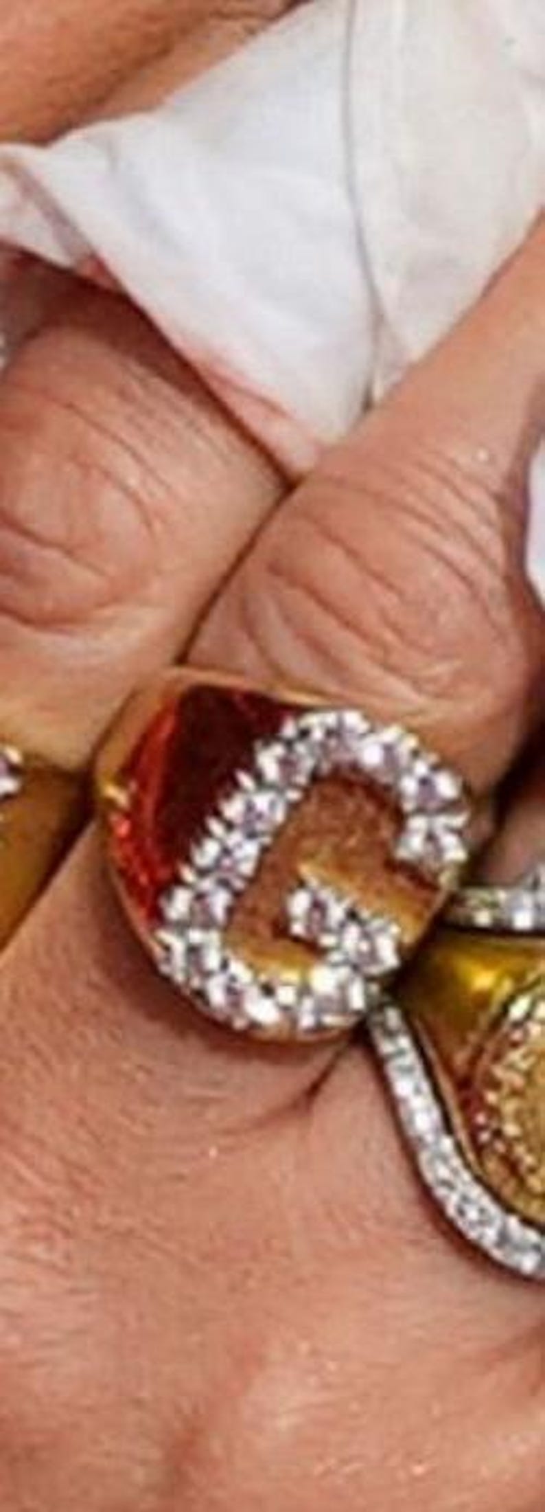 May include: Close-up of a gold-toned ring with a large, ornate design. The ring features a prominent letter "G" encrusted with small, sparkling crystals. The band and surrounding details are also gold.