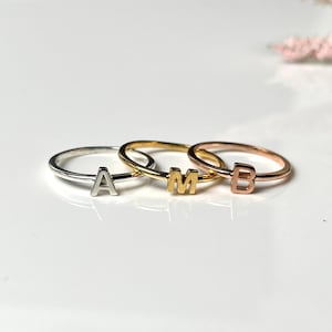Peut inclure: Trois bagues fines en métal disposées en rangée. La première bague est argentée avec la lettre A, la deuxième est dorée avec la lettre M et la troisième est en or rose avec la lettre B. Chaque bague comporte une seule lettre majuscule.