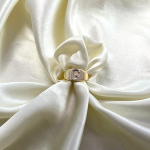 May include: A gold-colored ring with the letter "G" encrusted with small, clear gemstones. The ring is set against a backdrop of draped, off-white satin fabric, creating a luxurious aesthetic. The ring's band is smooth and polished.
