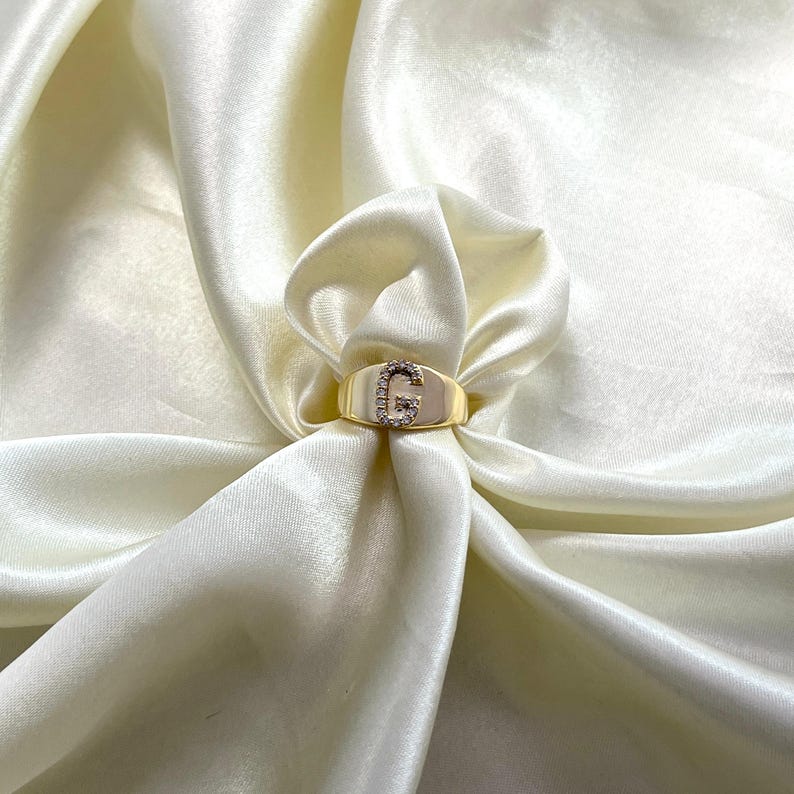 May include: A gold-colored ring with the letter "G" encrusted with small, clear gemstones. The ring is set against a backdrop of draped, off-white satin fabric, creating a luxurious aesthetic. The ring's band is smooth and polished.