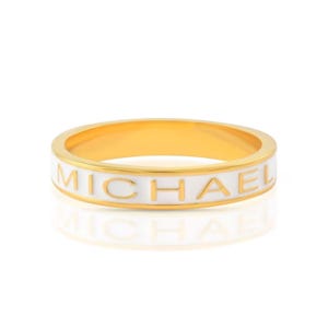 Puede incluir: Un anillo de oro con una banda de esmalte blanco que dice "MICHAEL" en letras doradas.