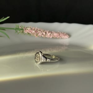 Pode incluir: Um anel de sinete de prata com uma parte superior ovalada elevada que apresenta a letra "A" gravada em uma fonte de escrita.