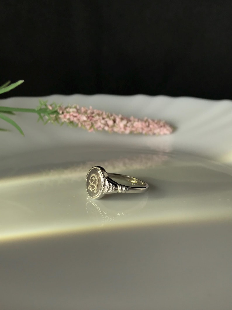 Pode incluir: Um anel de sinete de prata com uma parte superior ovalada elevada que apresenta a letra "A" gravada em uma fonte de escrita.