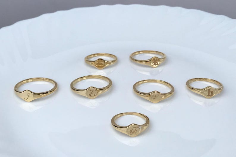 May include: Six gold signet rings with different initial letters engraved on the top. The rings are arranged in a semi-circle on a white surface.