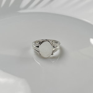 Può includere: Un anello d'argento con una pietra centrale ovale. L'anello ha una fascia decorativa con dettagli intricati. L'anello è appoggiato su una superficie bianca.