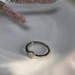 May include: A silver ring with a rounded oval top featuring the letter "S" engraved on the front.