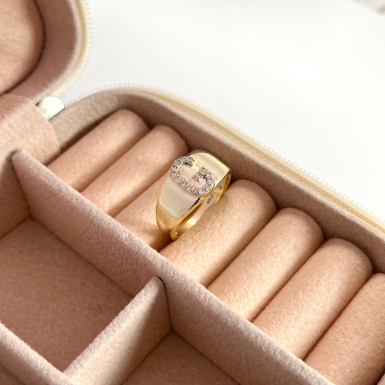 May include: A gold-colored ring with a white enamel band and a sparkling, gem-studded letter "G" design. The ring is set in a pink jewelry box, showcasing its elegant design. The ring is a piece of jewelry.