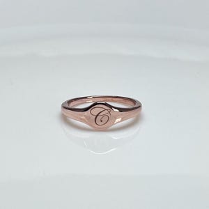 May include: A rose gold signet ring with a raised oval top featuring the letter "C" engraved in a script font.