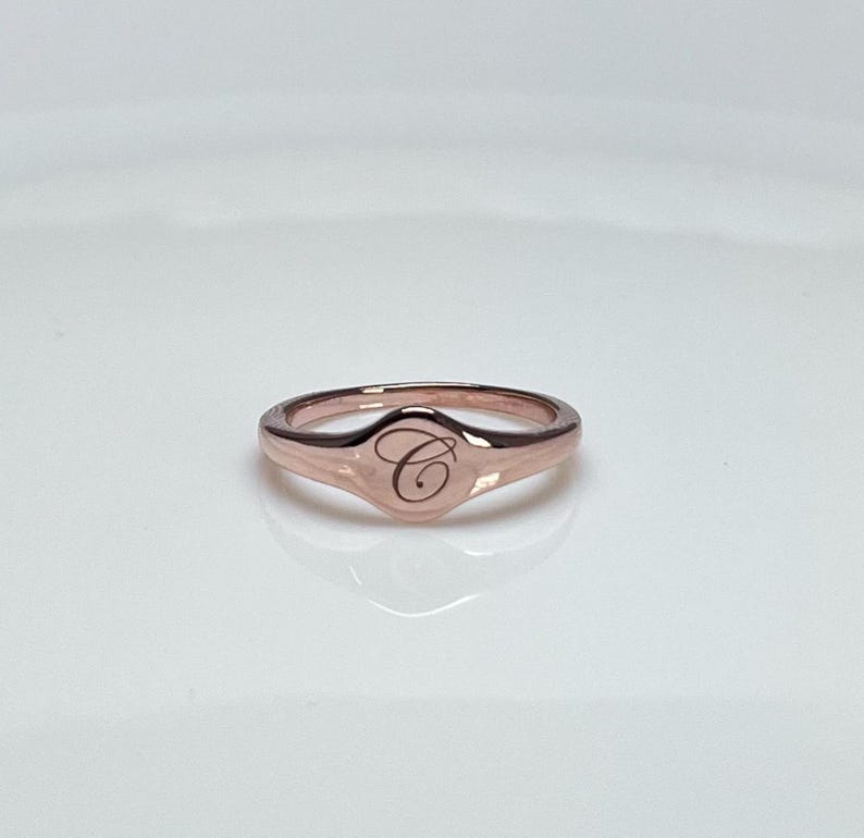May include: A rose gold signet ring with a raised oval top featuring the letter "C" engraved in a script font.