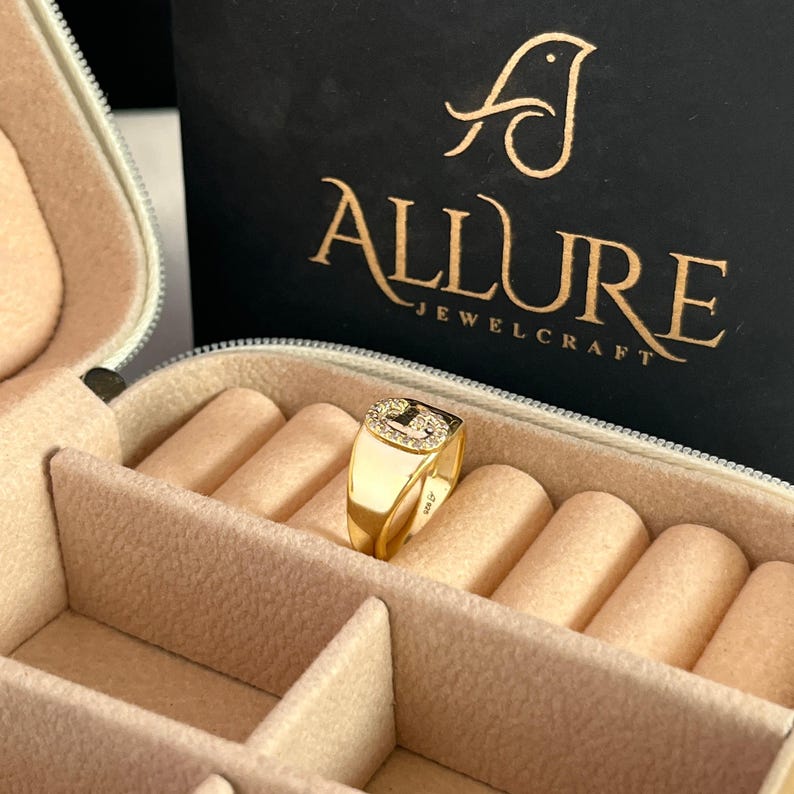 May include: A gold signet ring with a square face, set with small, clear stones. The ring is displayed in a beige jewelry box. The brand name "Allure Jewelcraft" is visible in gold lettering on a black background.
