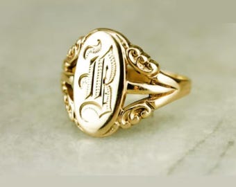 May include: A gold ring with an oval-shaped setting featuring a large, engraved letter "P". The ring band is decorated with intricate scrollwork.