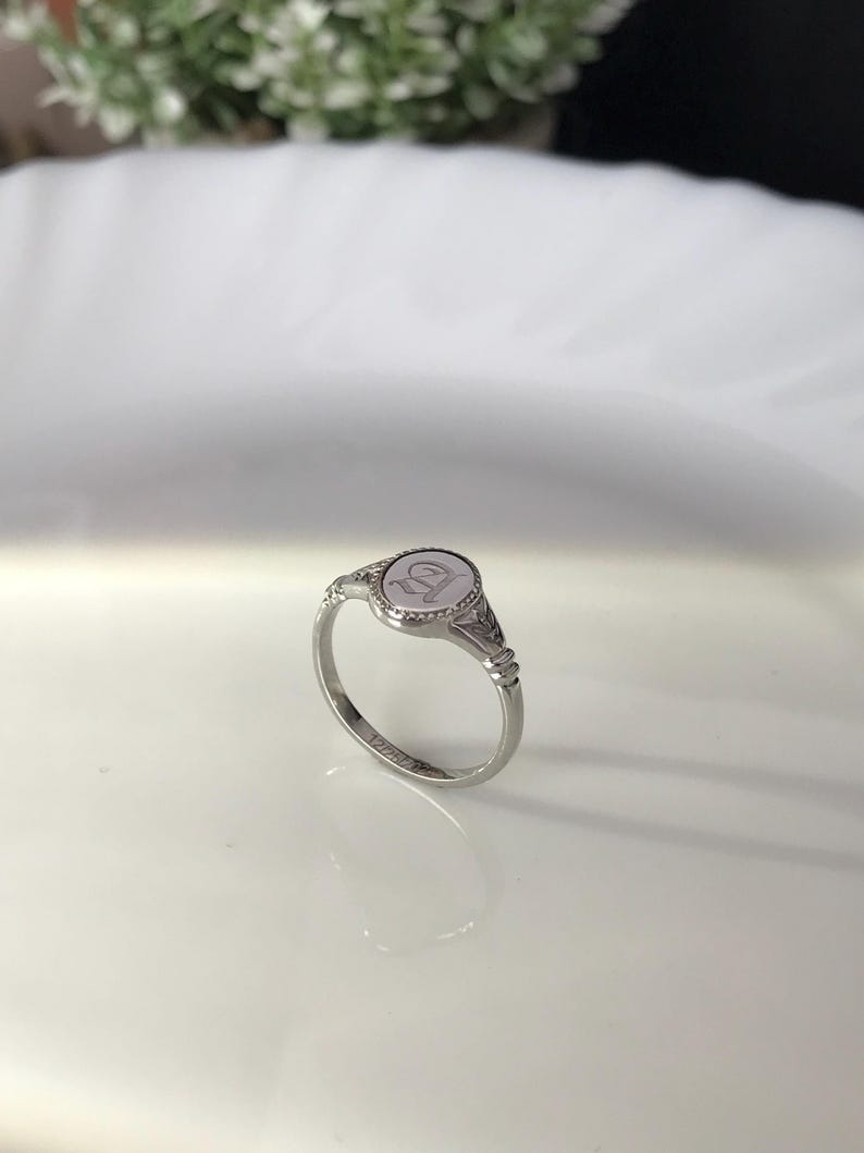 Pode incluir: Um anel de sinete de prata com uma parte superior ovalada em relevo que apresenta a letra "D".