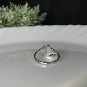 Pode incluir: Um anel de sinete de prata com uma parte superior lisa de forma oval. O anel est&aacute; sobre um prato branco.