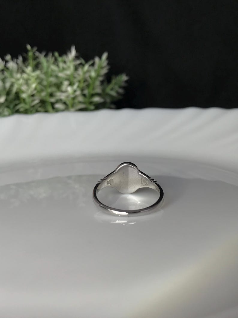 Pode incluir: Um anel de sinete de prata com uma parte superior lisa de forma oval. O anel est&aacute; sobre um prato branco.