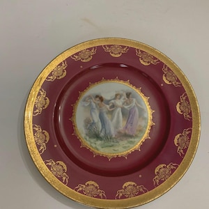May include: A decorative plate with a burgundy background and a gold rim. The center features a painting of women in flowing gowns. Gold floral designs adorn the rim. A vintage piece for display.