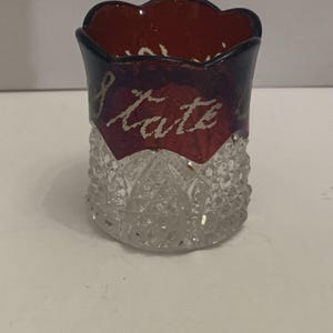 May include: A small, clear glass with a diamond-cut pattern on the lower half. The top rim is a deep red with a scalloped edge. The word "Tarte" is written in a cursive font on the side of the glass.