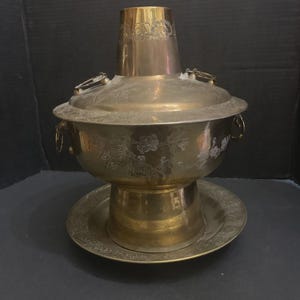 Puede incluir: Una olla caliente de latón vintage con tapa y asas, apoyada sobre una base a juego. La olla presenta diseños florales grabados y una chimenea cilíndrica alta. El diseño general es ornamentado y tradicional.