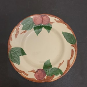 May include: A cream-colored ceramic plate with a decorative border featuring pink fruit, green leaves, and brown branches. The plate has a slightly scalloped edge and a smooth, glossy finish. A charming piece for serving or display.