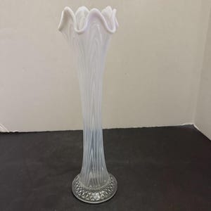 Könnte beinhalten: Eine hohe, weiße Glasvase mit gewelltem Rand. Die Vase hat einen schlanken, länglichen Körper mit vertikalen, strukturierten Linien. Der klare Sockel hat ein dekoratives, geometrisches Muster. Geeignet für Blumen.