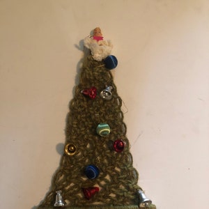 May include: A green crocheted Christmas tree wall hanging with a fringe bottom. The tree is decorated with small bells, buttons, and a small angel figurine at the top.