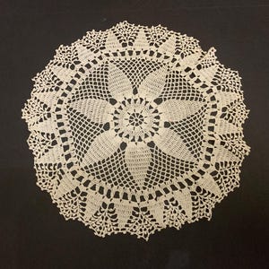 Puede incluir: Tapete de ganchillo hecho a mano de color crema con un diseño floral. El tapete circular presenta un motivo floral central rodeado de intrincados patrones calados y un borde festoneado. La delicada textura de encaje es visible sobre el fondo oscuro.