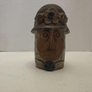 May include: A brown ceramic sculpture of a face wearing a hat. The hat is decorated with a flower and leaves. The face has carved features and a small, dark blue button at the base. A unique art piece.