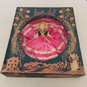 Puede incluir: Muñeca "dream doll" vintage en caja original. La muñeca tiene cabello rubio y viste un vestido rosa con patrones de estrellas y rayas. La caja turquesa presenta una ilustración de paisaje rosa y dorado y las palabras "dream doll".