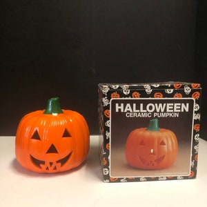 May include: A ceramic pumpkin with a carved face and a green stem. The pumpkin is in its original box, which is black with a white skull pattern and the text "HALLOWEEN CERAMIC PUMPKIN".