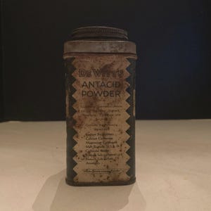 May include: A vintage rectangular metal tin for DeWitt's Antacid Powder with a weathered, dark finish and a silver-toned lid. The front features a serrated white border framing the product name and a list of ingredients printed in black text.
