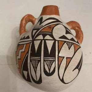 Puede incluir: Una pequeña jarra de cerámica pintada a mano con dos asas. La jarra está decorada con patrones geométricos en negro, naranja y blanco.