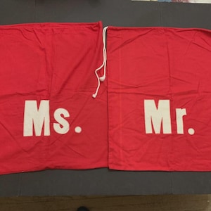 May include: Two red drawstring bags with white text. One bag reads "Ms." and the other reads "Mr.". The bags are made of a fabric material and have white drawstrings at the top.