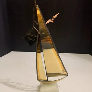 Puede incluir: Una escultura de velero de metal dorado con una base de mármol blanco. El velero tiene una pequeña bandera en la parte superior y un pájaro volando por encima. La escultura está firmada "Chery" y tiene una etiqueta que dice "Marie Jean Original".