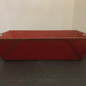 Puede incluir: Un contenedor rectangular de metal rojo con lados angulados y pequeños agujeros cerca de los bordes superiores. El contenedor tiene una apariencia ligeramente desgastada, lo que sugiere edad o uso. Se encuentra sobre una superficie oscura.