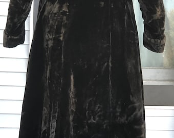 May include: A long, dark brown velvet coat with long sleeves and a flared skirt. The coat has a classic design and appears to be vintage. The fabric has a soft, textured appearance. The coat is worn over blue and gray socks.