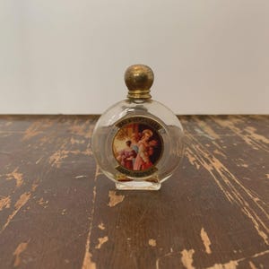 May include: A small, clear glass perfume bottle with a round body and a gold-colored cap. The bottle features a circular label with an illustration of people and the text "BAL A VERSAILLES" and "JEAN DESPREZ".