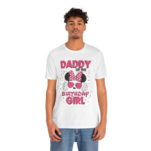 May include: White t-shirt with a Minnie Mouse-themed graphic. The design includes a pink polka-dot bow, sunglasses, and the text "DADDY OF THE BIRTHDAY GIRL" in pink. The shirt is worn by a person.