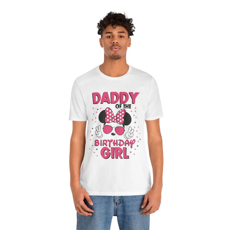 May include: White t-shirt with a Minnie Mouse-themed graphic. The design includes a pink polka-dot bow, sunglasses, and the text "DADDY OF THE BIRTHDAY GIRL" in pink. The shirt is worn by a person.