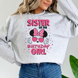 May include: A pale grey sweatshirt featuring the text "SISTER OF THE BIRTHDAY GIRL" in pink. The design includes a Minnie Mouse graphic with a pink bow, sunglasses, and peace signs.