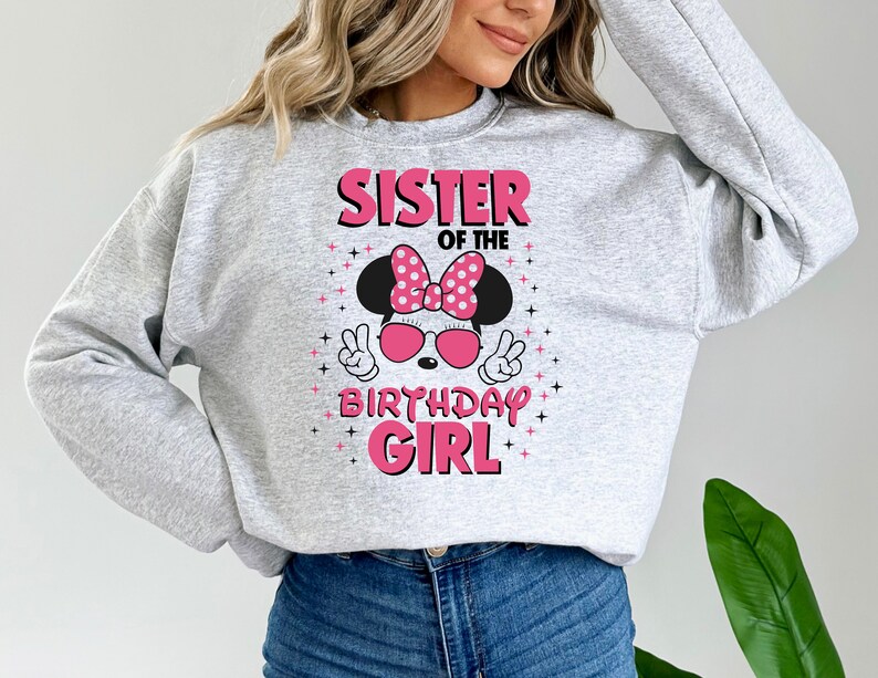 May include: A pale grey sweatshirt featuring the text "SISTER OF THE BIRTHDAY GIRL" in pink. The design includes a Minnie Mouse graphic with a pink bow, sunglasses, and peace signs.