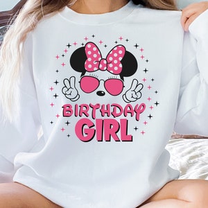 May include: White sweatshirt with a cartoon mouse wearing a pink polka-dot bow, sunglasses, and making peace signs. The text "BIRTHDAY GIRL" is in pink. Black and pink star accents surround the graphic. A casual, celebratory design.