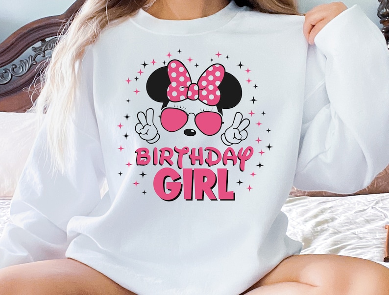 May include: White sweatshirt with a cartoon mouse wearing a pink polka-dot bow, sunglasses, and making peace signs. The text "BIRTHDAY GIRL" is in pink. Black and pink star accents surround the graphic. A casual, celebratory design.