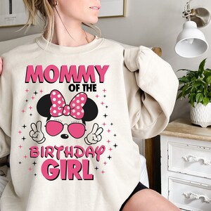May include: A beige sweatshirt with the text "MOMMY OF THE BIRTHDAY GIRL" in pink. The design features a cartoon character with a pink bow, sunglasses, and peace signs. Pink stars are scattered around the design.