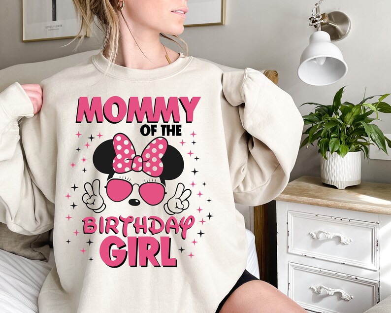 May include: A beige sweatshirt with the text "MOMMY OF THE BIRTHDAY GIRL" in pink. The design features a cartoon character with a pink bow, sunglasses, and peace signs. Pink stars are scattered around the design.