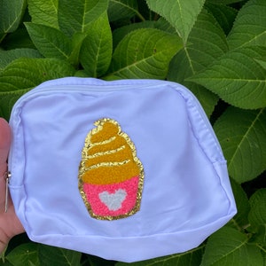 May include: A white zippered pouch with a gold and pink cupcake design. The cupcake has a gold swirl and a pink heart with a white center.
