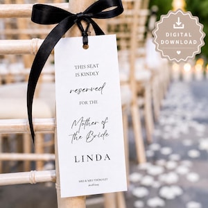 May include: A white wedding seat sign with black text and a black ribbon. The sign reads "THIS SEAT IS KINDLY reserved FOR THE Mother of the Bride LINDA". The sign is attached to a wooden chair.