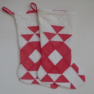 May include: Two quilted Christmas stockings with a red and white geometric pattern. The stockings have a diamond and triangle design, with a loop for hanging. The overall color scheme is red and white.