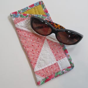 May include: Black sunglasses with brown lenses rest on a quilted glasses case. The case features a pink floral pattern with white triangles and a yellow interior. The case is trimmed with a floral border.