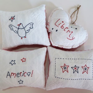 May include: Four white fabric pillows with red and blue embroidery. One pillow has a bird design, one has the word "Liberty", one has the word "America!" and one has three stars.